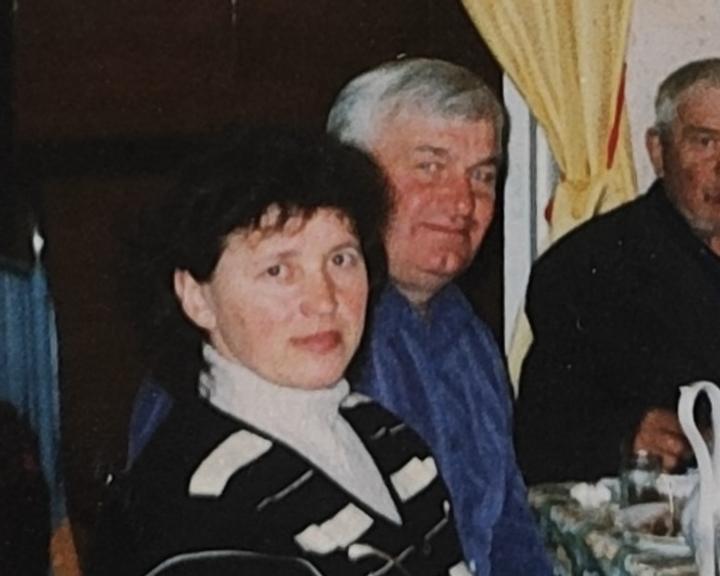 Жінка в смугастому светрі з білим комірцем і сивий чоловік у синій сорочці сидять за святковим столом, позаду жовті штори