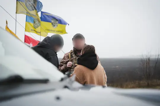 Журналістка спілкується з військовими поруч зі стелою. За нею — просторе поле, куди ступати небезпечно.
