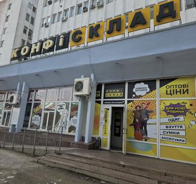 Фасад магазину «Конфі Склад» у Сумах: велика жовто-чорна вивіска, вітрини з написами «Оптові ціни» та «Одяг, взуття, сумки, аксесуари»