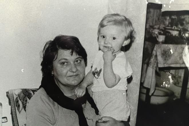 Юлія Опанасенко з бабусею Ольгою. Пізніше саме бабуся навчила її плавати Чорно-біле архівне фото: жінка з короткою стрижкою та темним шарфом тримає на руках світловолосого малюка в кімнаті сільської хати