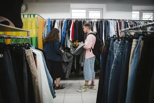 Дівчина в джинсовій куртці й хлопець з наплічником переглядають одяг на вішаках у секонд-хенді в Сумах