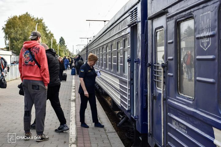 На платформі біля синього пасажирського вагона провідниця витирає борт ганчіркою, поруч стоять пасажири з валізами.