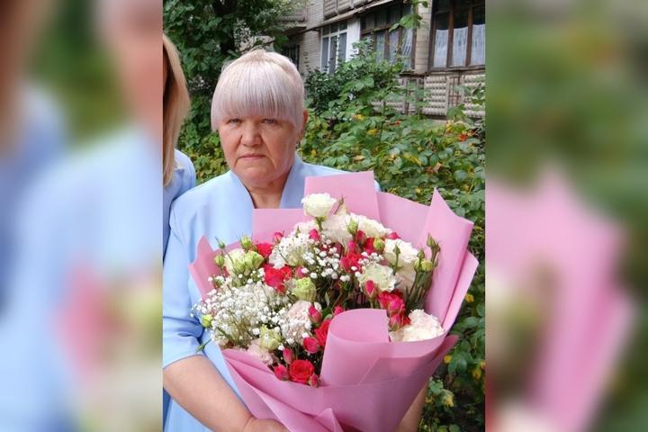 Літня жінка з світлим волоссям у блакитній сукні тримає букет троянд і гіпсофіли біля багатоповерхівки
