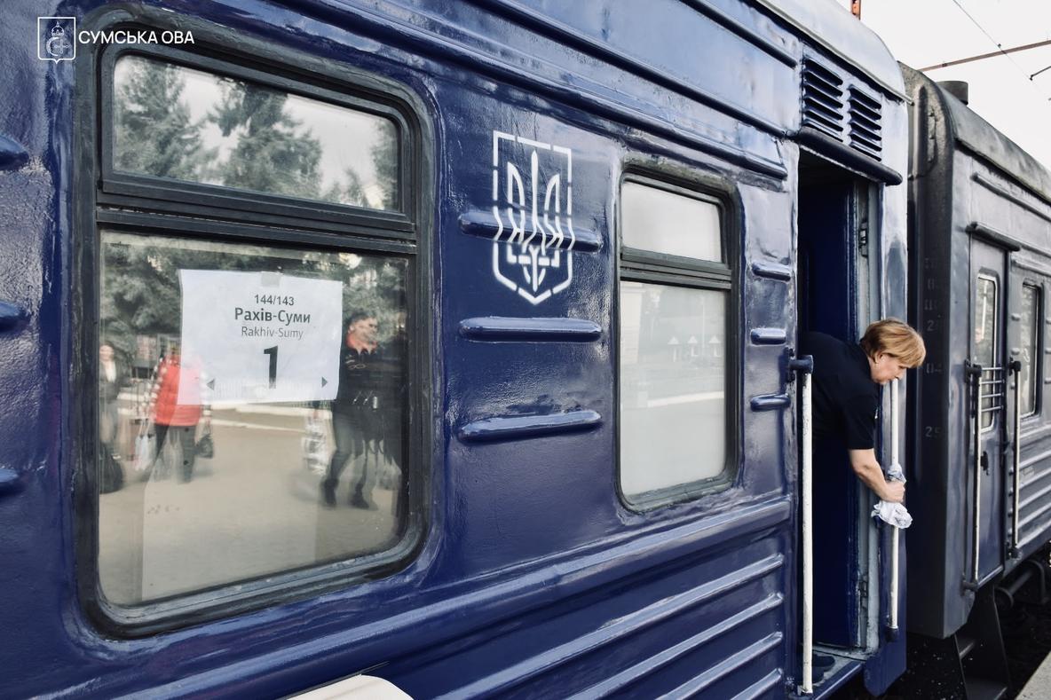 Темно-синій вагон із табличкою «144/143 Рахів — Суми, 1» на склі; у дверях провідниця тримається за поручень із ганчіркою.
