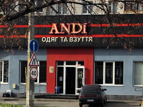 Магазин ANDI «Одяг та взуття» у Сумах з червоною вивіскою, пандус із поруччям біля входу, дорожні знаки на стовпі