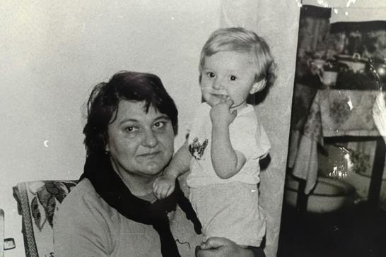 Юлія Опанасенко з бабусею Ольгою. Пізніше саме бабуся навчила її плавати Чорно-біле архівне фото: жінка з короткою стрижкою та темним шарфом тримає на руках світловолосого малюка в кімнаті сільської хати