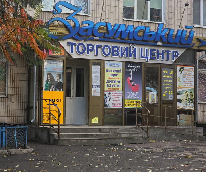 Вхід до ТЦ «Засумський» у Сумах: синя вивіска, двері з вивісками магазинів дитячого, військового одягу, взуття й сумок, сходи з поруччям
